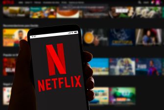 Close-up of a hand holding a smartphone displaying the Netflix logo, with a blurred Netflix streaming interface in the background.