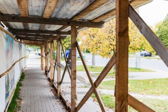 Sidewalk shed structure covering a walkway with wooden supports and a roof, surrounded by autumn foliage and parked cars in the background.