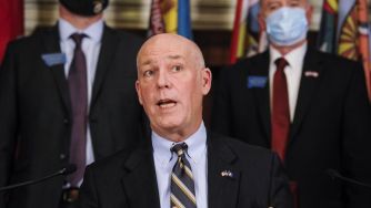 A man in a suit speaks at a press conference, with two individuals in the background, one wearing a mask. The setting includes flags hanging on the wall.