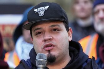Close-up of a young man wearing a black cap with a bull logo, speaking into a microphone at a public gathering with a blurred crowd in the background.