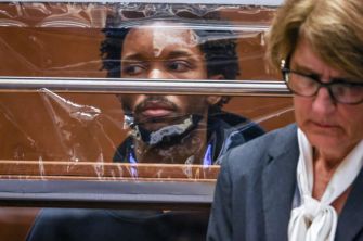 Young man sitting in a courtroom behind a glass barrier, looking to his left, with a lawyer nearby.