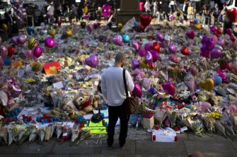 A person stands in front of a large memorial made up of flowers, balloons, and messages honoring the victims of the 2017 Manchester concert bombing.