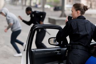 Police officers in pursuit of a suspect, one using a radio while the other chases a fleeing individual.