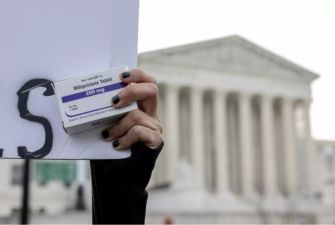 Close-up of a hand holding a box labeled Mifepristone (200 mg) beside a protest sign, with the U.S. Supreme Court building in the background.