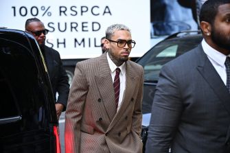 Man with light hair and beard wearing a brown plaid suit, white shirt, and burgundy tie, sunglasses, walking outdoors beside a black car.