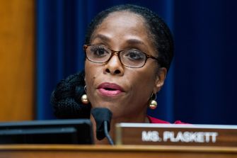 Close-up of a woman wearing glasses speaking into a microphone at a congressional hearing, with a nameplate in front.