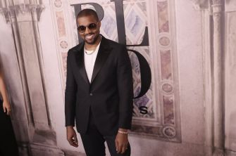 Man in a black suit, white shirt, sunglasses, and gold chain posing for a photo at a formal event backdrop.