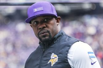 Close-up of a man wearing a purple Minnesota Vikings cap and jacket, looking to the left on a football field.