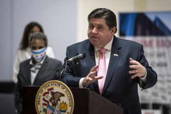 Governor J.B. Pritzker speaking at a press event regarding Illinois' efforts to end federal immigration detention programs, with a person in the background.