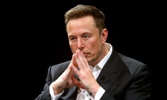 Elon Musk seated with his hands clasped in front of his face, looking serious.