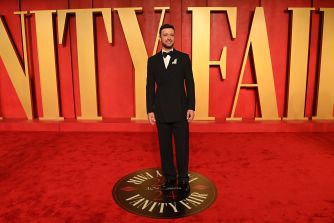 Man in a black tuxedo posing on a red carpet at a Vanity Fair event, with large gold Vanity Fair letters in the background.