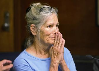 A woman with gray hair and glasses, appearing emotional with her hands clasped in front of her mouth, sits in a courtroom setting.