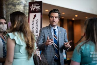 Man in a light blue blazer and striped tie speaks to a group of people in a hallway.