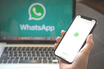 A person holding a smartphone displaying the WhatsApp logo, with a laptop in the background also showing the WhatsApp logo.