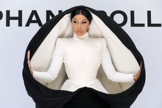 Woman wearing a white high-neck dress, with a black cape opened around her arms, posing in front of a backdrop.