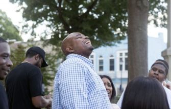 A man joyfully laughing while surrounded by friends and family in a sunny outdoor setting.
