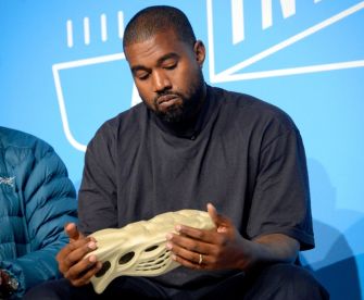 Man in a dark shirt sitting and examining a white foam sneaker against a blue backdrop.