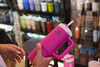 Close-up of a pink Stanley tumbler with a white straw being held by a person in a store, with blurred shelves in the background.