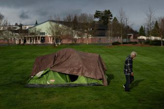 A person walks past a tent set up on a grassy area, indicating the presence of homelessness.