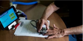 Close-up of an elderly person's hand and a caregiver's gloved hand over a desk, with a pulse oximeter displaying Blood Oxygen and papers on the table.