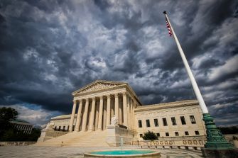 The U.S. Supreme Court building with its columned façade, a tall flagpole displaying the American flag, set against a dark, stormy sky.