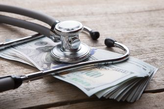 A stethoscope resting on a pile of hundred-dollar bills on a wooden surface.