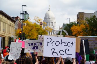 Pro-choice demonstration with people holding signs, including “Protect Roe,” in front of a dome-topped capitol building and autumn trees.