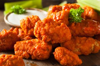 Close-up of crispy orange-buffalo style chicken bites piled on a wooden surface with a small dish of dipping sauce and parsley garnish.