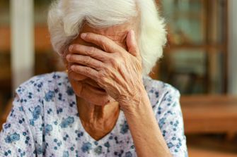 An elderly woman with white hair covering her face with one hand, expressing distress or concern.