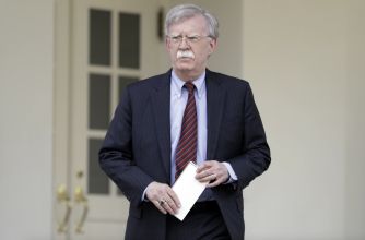 Older man with gray hair and mustache, wearing a dark pinstripe suit and red striped tie, holding a white envelope while walking outside a light-colored building.