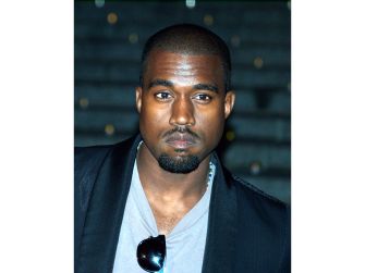 Portrait of a man with short hair and a goatee wearing a light gray shirt and dark jacket, sunglasses tucked at the neckline, looking at the camera.