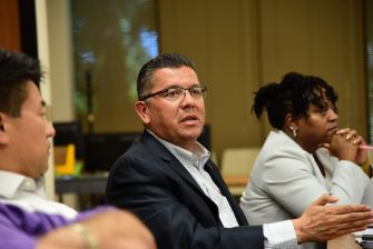A man in a dark suit and glasses speaks at a table in a conference room, with a woman seated in the background.