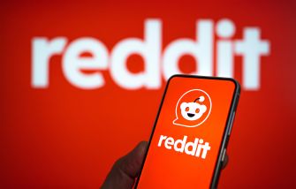 Close-up of a smartphone showing the Reddit app icon, held in a hand against a red background featuring the Reddit logo.