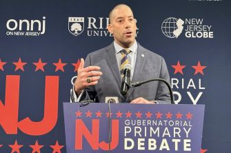 A man speaking at a gubernatorial primary debate podium with a background featuring political branding and logos.