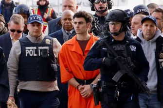 Suspect Luigi Mangione in handcuffs, wearing an orange jumpsuit, surrounded by law enforcement officers during his court appearance.
