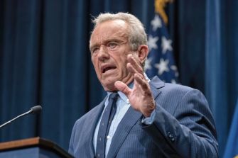 Older man in a blue pinstripe suit speaking at a podium, gesturing with his hand, with an American flag in the background.