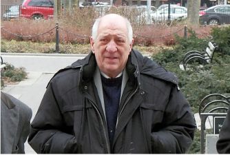 Elderly man in a black coat walking in a park setting, surrounded by trees and city buildings.