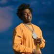 Rapper Kodak Black standing with a chain and microphone, wearing an orange sweatshirt.