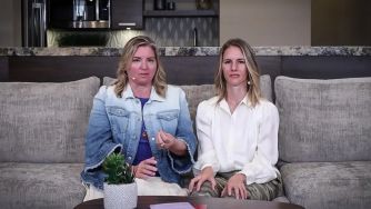Two women seated on a couch, discussing their work or experiences, with a plant and table in front of them.