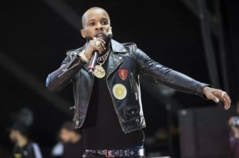 Tory Lanez performing on stage, wearing a black leather jacket and holding a microphone.