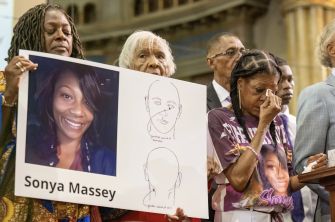Mourners at a vigil holding large signs with a woman’s photograph and drawings, many appearing emotional.