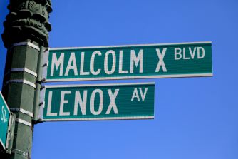 Street signs reading Malcolm X Blvd and Lenox Ave in New York City, shown against a clear blue sky.