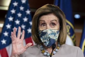 A woman wearing a mask gestures while speaking at a podium with flags in the background.