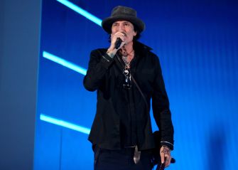 Male rock singer wearing a black outfit and hat, singing into a microphone on stage with blue lighting and visible tattoos.