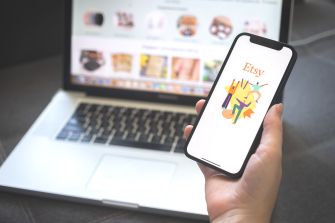 A person holding a smartphone displaying the Etsy logo, with a laptop in the background showing the Etsy store interface.