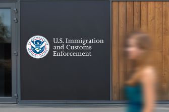 Doorway with the U.S. Immigration and Customs Enforcement logo on a dark panel and a blurred person walking past