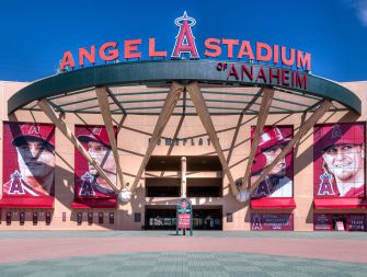 Front view of Angels Stadium of Anaheim entrance with a red-letter sign and player banners against a clear blue sky.