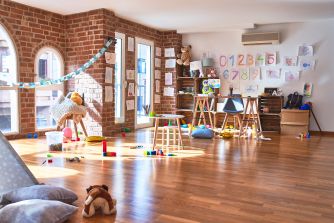 A brightly lit daycare room with wooden floors, child-sized furniture, educational posters on the walls, and scattered toys on the floor.