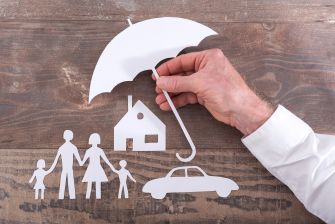 Hand holding an umbrella over paper cutouts of a family, a house, and a car, symbolizing insurance protection and coverage.