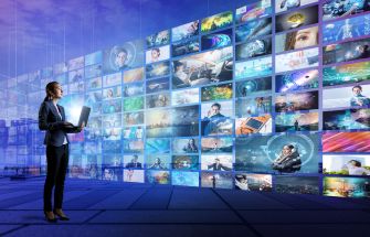A person holds a laptop in front of a large wall displaying multiple digital screens showcasing various video content.